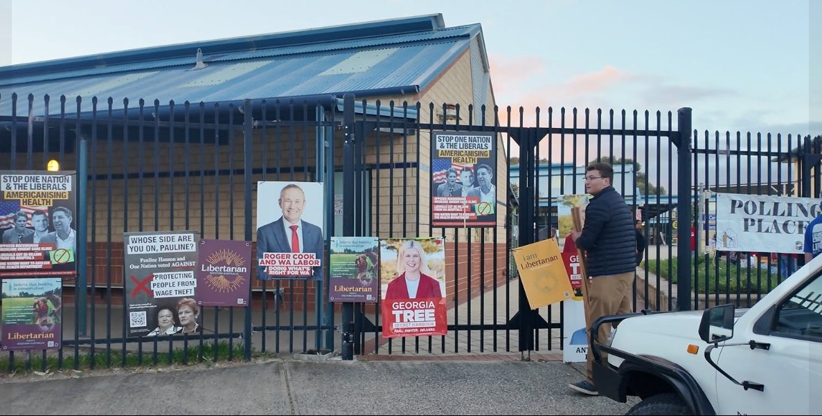 Corflutes from several parties zip-tied to the fence outside a polling place at dawn, with the polling place banner visible at the end of the fence.