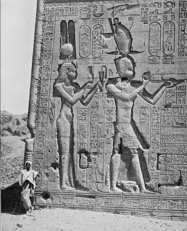 Early photograph of a relief on the south exterior wall at Dendera: two crowned figures making offerings, carved at several times life size, a man standing at the foot of the wall for scale.
