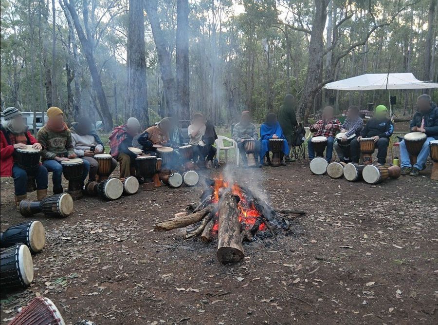 A wide circle of men seated on camp chairs around a burning log fire in open jarrah forest at dusk, each with a djembe drum, smoke rising through the trees. Faces are blurred.