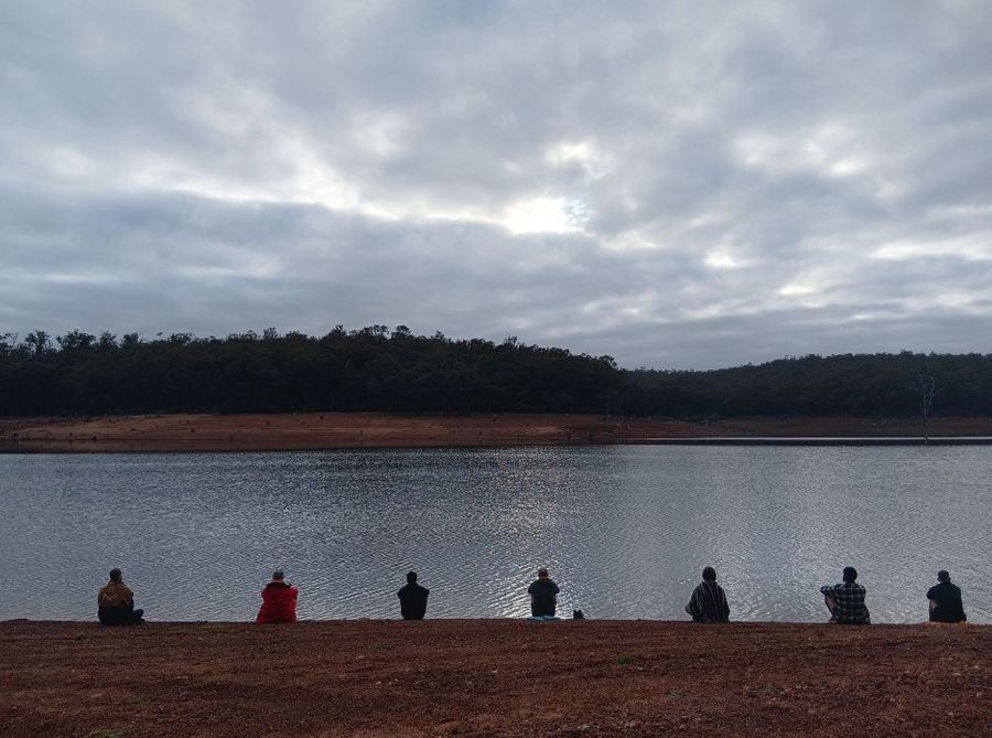 Seven men sitting apart along a red clay bank at the edge of still water, facing forest on the far side under heavy grey cloud in the early morning.
